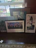 Four framed art prints grouped on floor near window. Includes a Key West lighthouse print, a Monterey Bay Aquarium sea otters print, a Jamie Wyeth print, and a partially obscured piece. Frames vary in size and need cleaning.