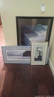 Three framed art prints, including a coastal landscape and two smaller images leaning against a wall, showing the size and framing style.