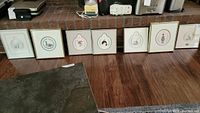 Photo showing 7 framed prints lined on the floor with seasonal animal ornament shapes inside, each framed with white matting and thin framing.