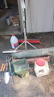 Overview of garden tools and equipment on ground outside, including handheld tools, seed spreader, broom, gravel rake, hose, and containers