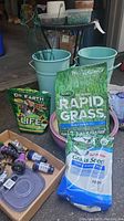 Gardening supplies assortment showing Scotts Rapid Grass fertilizer, Scotts Turf Builder Grass Seed, extra grass seed bag, green plastic pots, assorted hose nozzle attachments and fittings in a box.