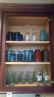Shelved assortment of glasses including dark blue, textured turquoise, red hand-blown juice glasses, clear wine stemware, and plastic tumblers.