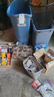 Overview of lot contents including 2 wastebaskets, glass canning jars, cardboard boxes with light bulbs and other items.
