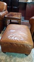 Photo showing a brown leather ottoman and a solid wood side table with a scratched top, positioned on a patterned rug between leather chairs.