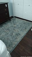 Light blue area rug with intricate floral and paisley pattern, shown on dark wood floor near a dresser and bed.