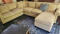 Wide angle photo showing the full L-shaped sectional sofa in beige with multiple seat and back cushions and blue accent pillow on the right chaise lounge section. The fabric is creamy beige and shows some wear.