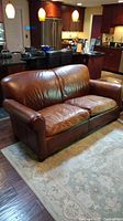 Front and right side view of a brown leather three-seat sofa in a modern kitchen and living area, showing leather texture on cushions and armrest.
