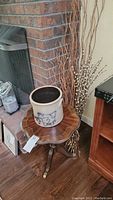 Wooden vintage side table with a scalloped edge and tripod base, stoneware pot, and decorative branches arranged together near a fireplace.
