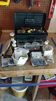 Black toolbox open on a workbench surrounded by various hardware items including screws, nails, safety glasses, and small containers.