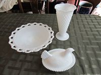 Three white milk glass pieces on a dark green checkered tablecloth: a pedestal cake stand with scalloped edge, a hobnail patterned vase, and a covered dish shaped like a chicken with a beaded rim.