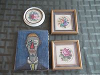 Four art pieces shown together on surface, three floral needlework pieces and one abstract portrait on canvas.