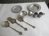 Overview of the lot showing assorted brass serving utensils and pewter plates and creamers on a white background.