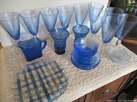 Vintage blue glass dishware set shown on a table including goblets, plates, pitchers, and small bowls made of different blue glass tones.