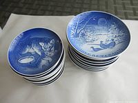 Two stacks of blue and white vintage plates, one stack smaller featuring cats, and another larger stack with winter and domestic scenes