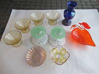 Full view of all 11 coloured glass decorative items arranged on a white background. Includes six amber dessert bowls, two floral hand-painted candlesticks with green tops, a cobalt blue vase, an orange heart-shaped bowl, and two small decorative plates.