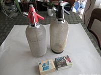 Two vintage glass syphon bottles with metal mesh coverings and two boxes of soda syphon chargers set on a table.