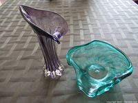 Purple glass vase with fluted trumpet shape and clear textured base beside teal freeform glass bowl on fabric surface.