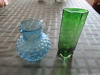 Full view of the green glass vase and blue hobnail pitcher placed on a checkered tablecloth.