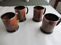 Four brown ceramic beer steins with black handles and bases displayed on white cloth, showing overall set