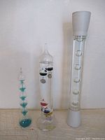 Three tall vintage glass instruments shown side by side on a surface against white wall background: Galileo thermometer, glass boiler, and lava lamp-like object.