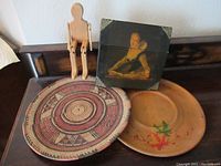 First photo showing woven basket, wooden figurine sitting on basket, framed portrait leaning on basket, wooden plate on table.
