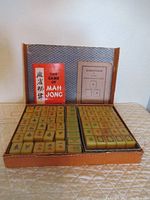Opened box displaying Mahjong tiles organized in rows, red booklet titled 'The Game of Mah Jong' and directions sheet inside box lid.