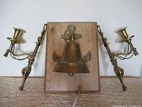 Full view of brass bell mounted on wood with pair of brass wall sconces on each side.