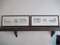 Pair of framed prints displayed on shelf depicting coastal and village scenes.