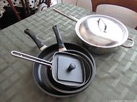 Four pans arranged on a green patterned table, featuring a large stainless steel Lagostina pan with lid and three non-stick pans with two black handles and one stainless handle.