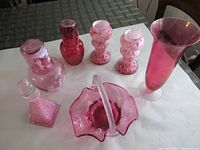 Full view of 7 pink glass items on table including vases, decanters, basket bowl and candle holders.