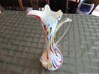 Tall handmade glass vase with multicolor swirls and clear handle shown from the side on a table.