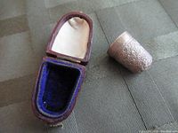 Open leather case with blue velvet and cream satin lining sitting on fabric surface with the metal thimble beside it.