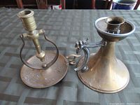 Two vintage brass candle holders shown on tabletop, one with round engraved base, one with bell-shaped base and animal head handle