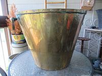 Full view of vintage brass kindling basket with handles visible, placed on a glass table indoors.