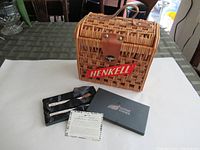 Photo of the wicker Henkell basket with leather strap and red HENKELL label sitting on a table, alongside the silver-plated pâté knife set in black packaging and an informational card.