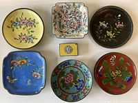 Top-down view of six cloisonné plates and dishes arranged flat showing floral patterns on various colored backgrounds including yellow, blue, red, and dark tones