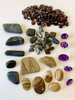 Wide view of the assortment of cabochons and faceted stones laid out on white surface showing groups of brown picture jasper, dark iridescent labradorite, deep red garnet, and purple amethyst stones.