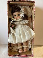 Photo of Ginny Vogue Doll in original box wearing white party dress and lace bonnet, showing her face, hair, and clothing details.
