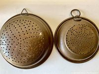 Two antique copper strainers side by side, showing tops with perforated holes and hanging loops