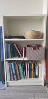 White wood bookshelf with three shelves containing books and two fabric storage baskets.