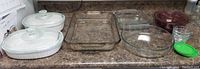 Overview of CorningWare white oval casserole dishes with lids on left, clear rectangular Pyrex baking dish, clear round Pyrex glass dish, glass mixing bowls with one burgundy, and small glass storage containers with green lid on counter