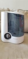 White Honeywell humidifier shown assembled on a beige couch with translucent water tank and rotary dial.