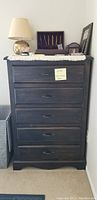 Front view of tall wooden dresser with five drawers and metal handles. Fabric runner on top with lamp and various decorative items (not included).