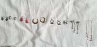 Photo of assorted 11 pairs of earrings laid out on white fabric background showing silver, gold-tone and costume jewelry in various styles including hoops, studs, and dangles.