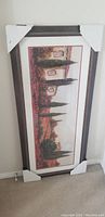 Photo of the framed Italian landscape picture leaning against a wall, with protective corner covers on the frame.