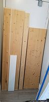 Five wood pieces leaned against a white wall. Four pieces are natural wood and one is white painted wood, all rectangular and flat.
