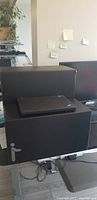 Front view showing black two-tier standing desk with laptop on lower step, monitor behind it on desk.