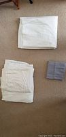 Folded white bedsheets and a small folded gray item (pillowcase) on carpeted floor, showing condition and color of sheets.