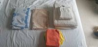 Photo showing three folded towels: blue/white patterned, beige, and white towels plus a stack of six microfiber cloths in various colors on a bed.
