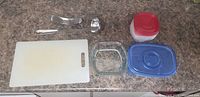 Top down view of Iron Clad soup ladle, garlic press, plastic containers with lids, Pyrex glass dish, and plastic cutting board on countertop